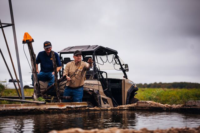 Two men sit in a utility vehicle parked beside a serene pond, surrounded by greenery and natural scenery.